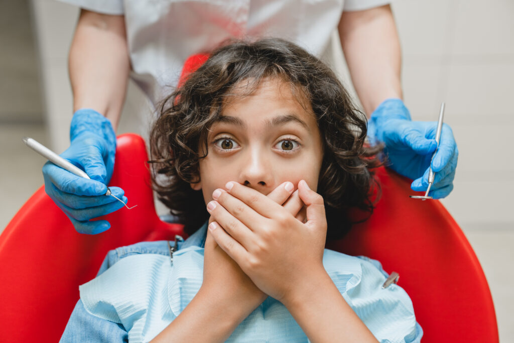 A young child sitting in a dental chair covering their mouth in fear during pediatric dental emergency treatment